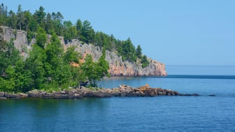 A forested rhyolite headland with salmon-pink rock faces juts into Lake Superior under a clear blue sky, with a low rocky shoreline in the foreground and calm blue water stretching to the horizon.